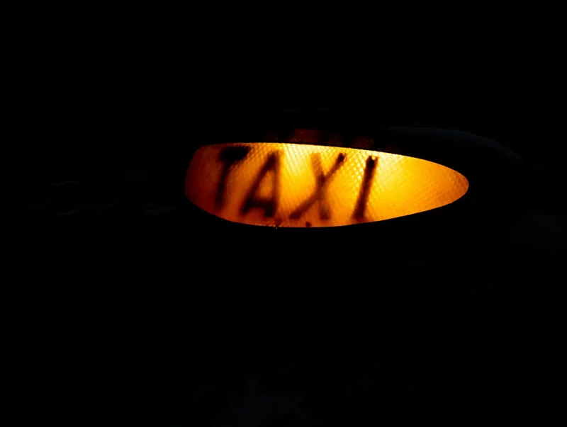 The orange TAXI sign on the roof of a black cab lit up in the dark, showing the cab is free to hail