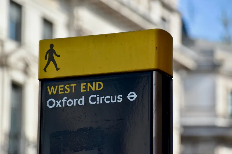 A yellow London walking sign with a pedestrian symbol pointing to the West End and Oxford Circus Underground station