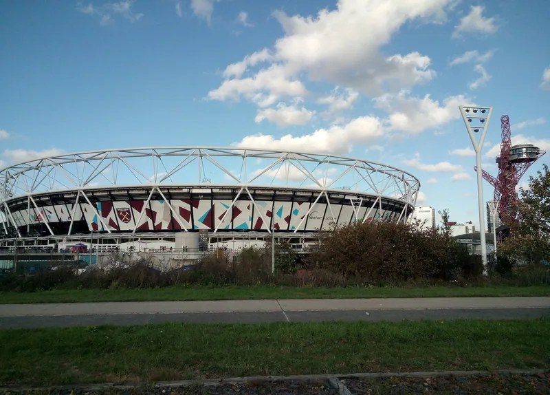 The London Stadium's white roof truss and angular cladding seen across scrub from the Greenway path, the Orbit tower to the right