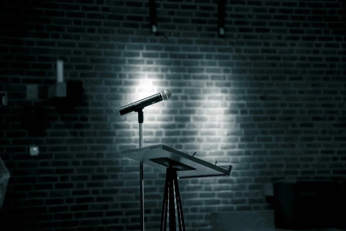 A microphone on a stand lit by a single spotlight against the bare brick wall of a club stage