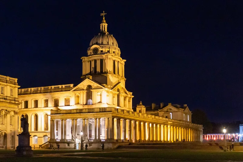 The Old Royal Naval College in Greenwich floodlit at night, with its domed tower above the lit colonnade
