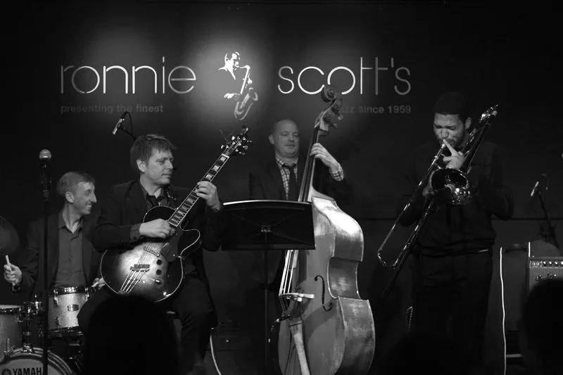 Black-and-white stage shot of four musicians with guitar, double bass, trombone and drums under the Ronnie Scott's backdrop