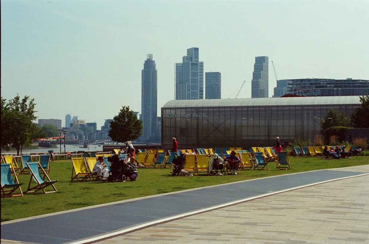 Rows of striped deckchairs on a riverside lawn with people sitting out, Thames-side towers behind