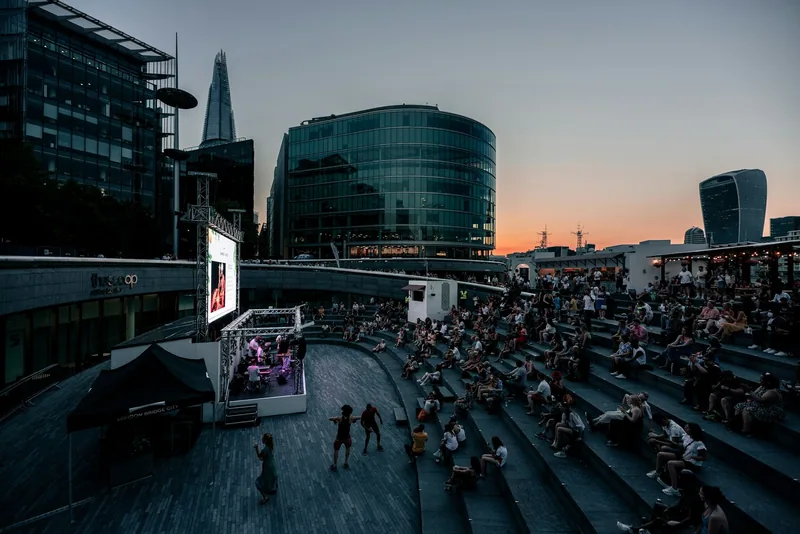 A crowd on the curved steps of a riverside amphitheatre watching a big screen on a warm summer evening
