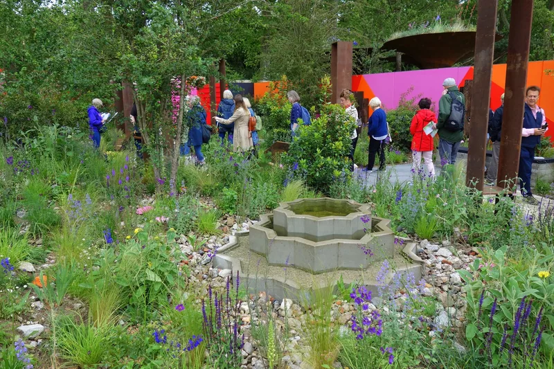 Visitors looking at a show garden of loose perennial planting around a stone water bowl at the Chelsea Flower Show