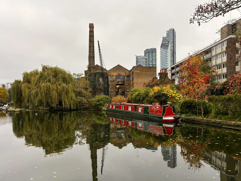 A red narrowboat moored on a London canal below autumn willows, with tower blocks behind