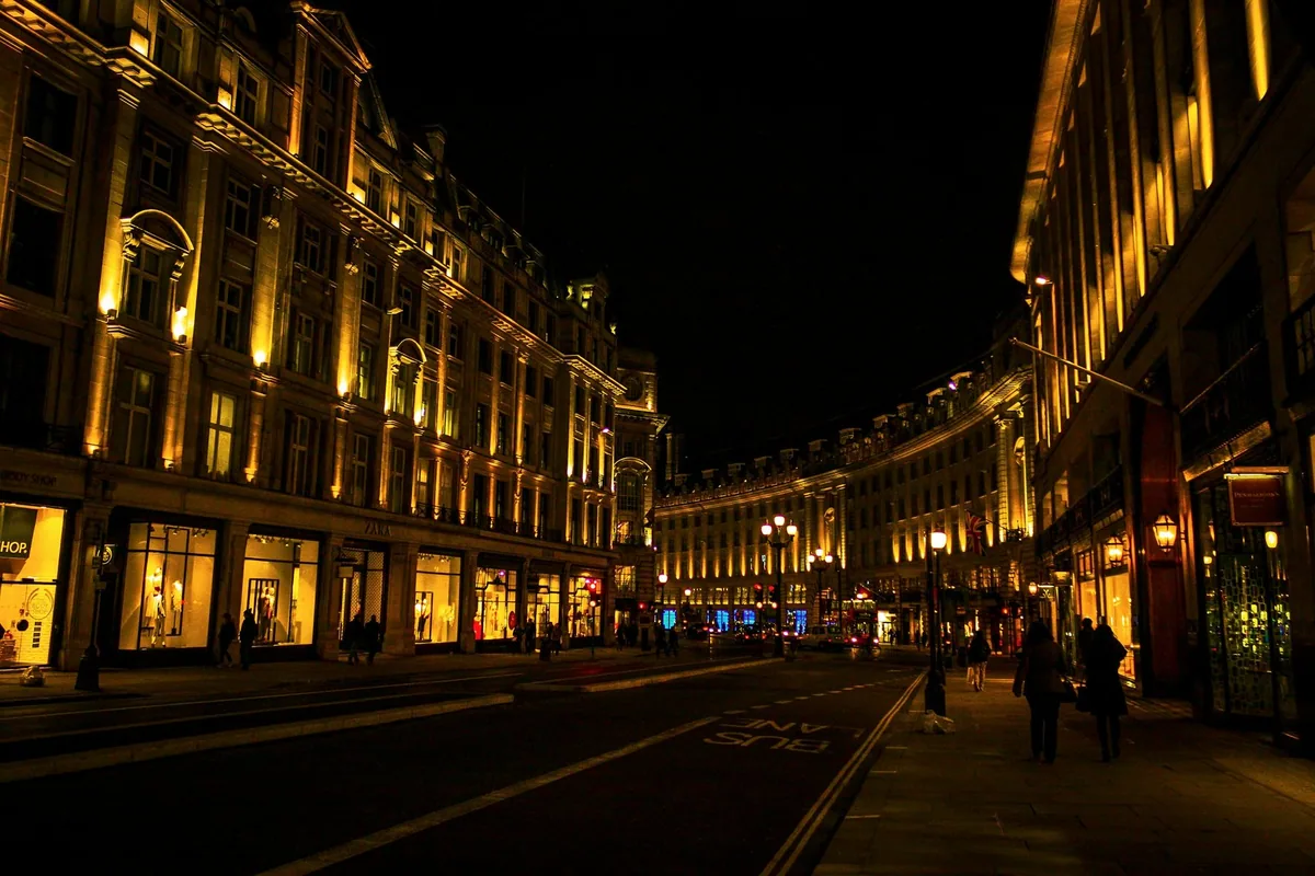 People walking past golden-lit shopfronts along a curving London street after dark