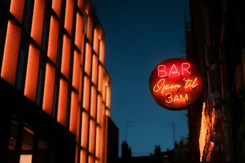 A red neon sign reading Bar, open til 3am, glowing on a building wall at night