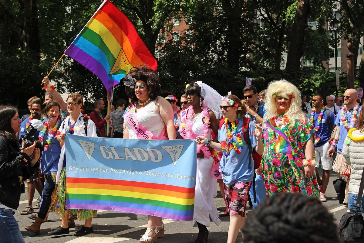 Marchers carrying a rainbow banner and flag along a tree-lined London street during the Pride parade