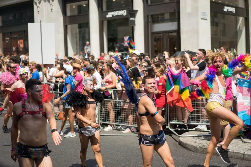 Dancers in the road at Pride in London, with spectators waving rainbow flags behind crash barriers