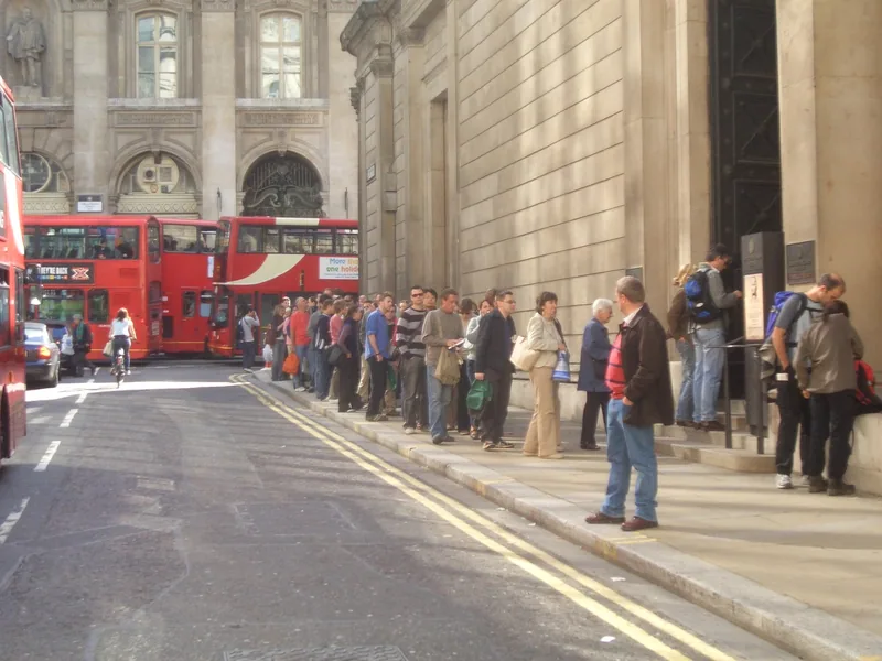 People queuing beside the stone wall of the Bank of England, with red buses passing in the street