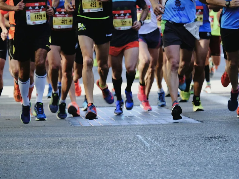 A tight pack of runners with numbered race bibs crossing a timing mat on a closed road, photographed from knee height