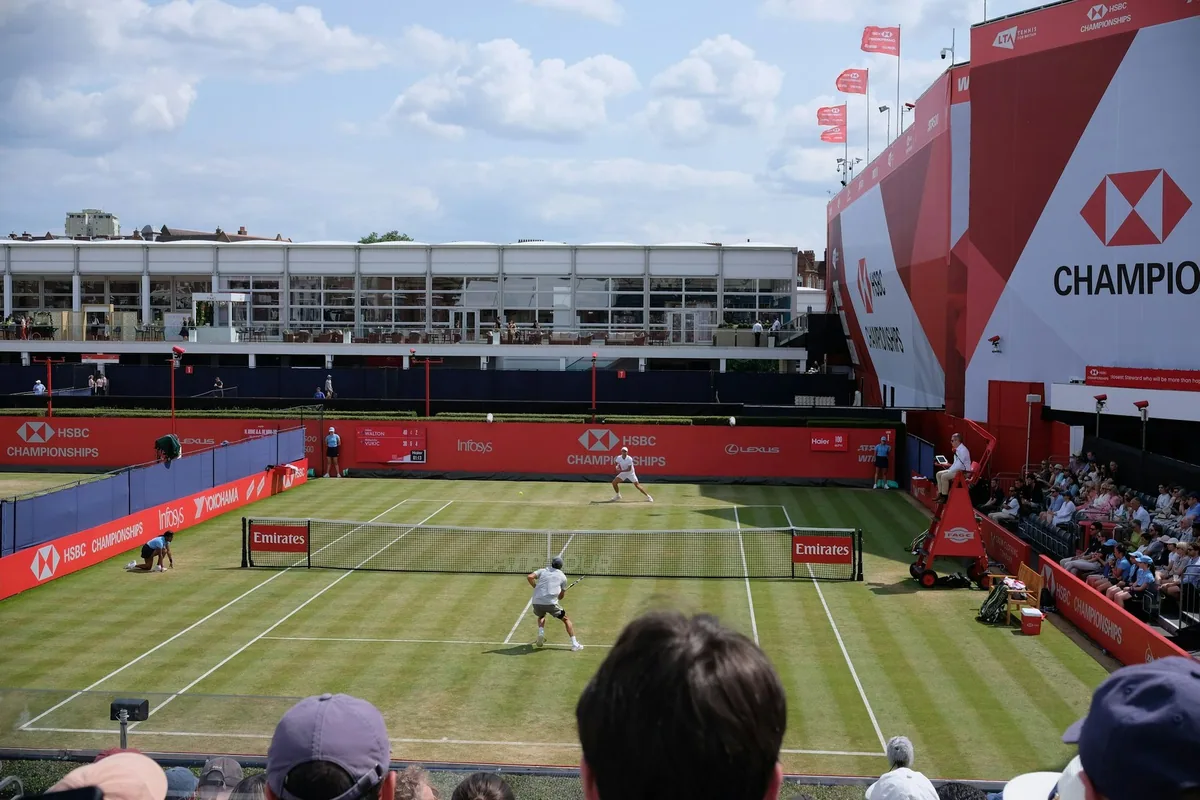 A grass-court match in progress at the Queen's Club HSBC Championships, seen over the heads of spectators, with the clubhouse balcony behind