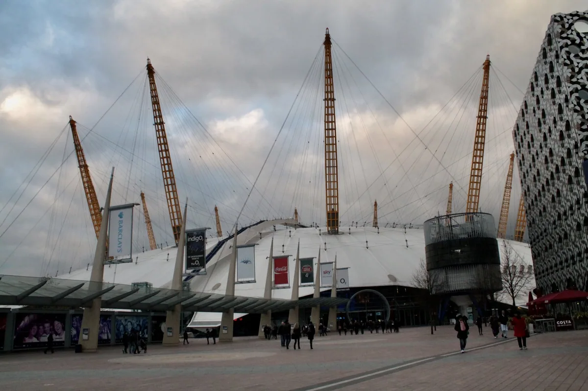 The tented roof and yellow masts of the O2 rising above the entrance square, with people walking past
