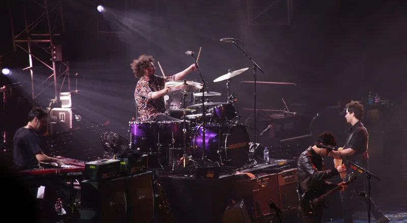A band on stage at the O2 Arena, the drummer raised on a riser between keyboards and guitars in white spotlights