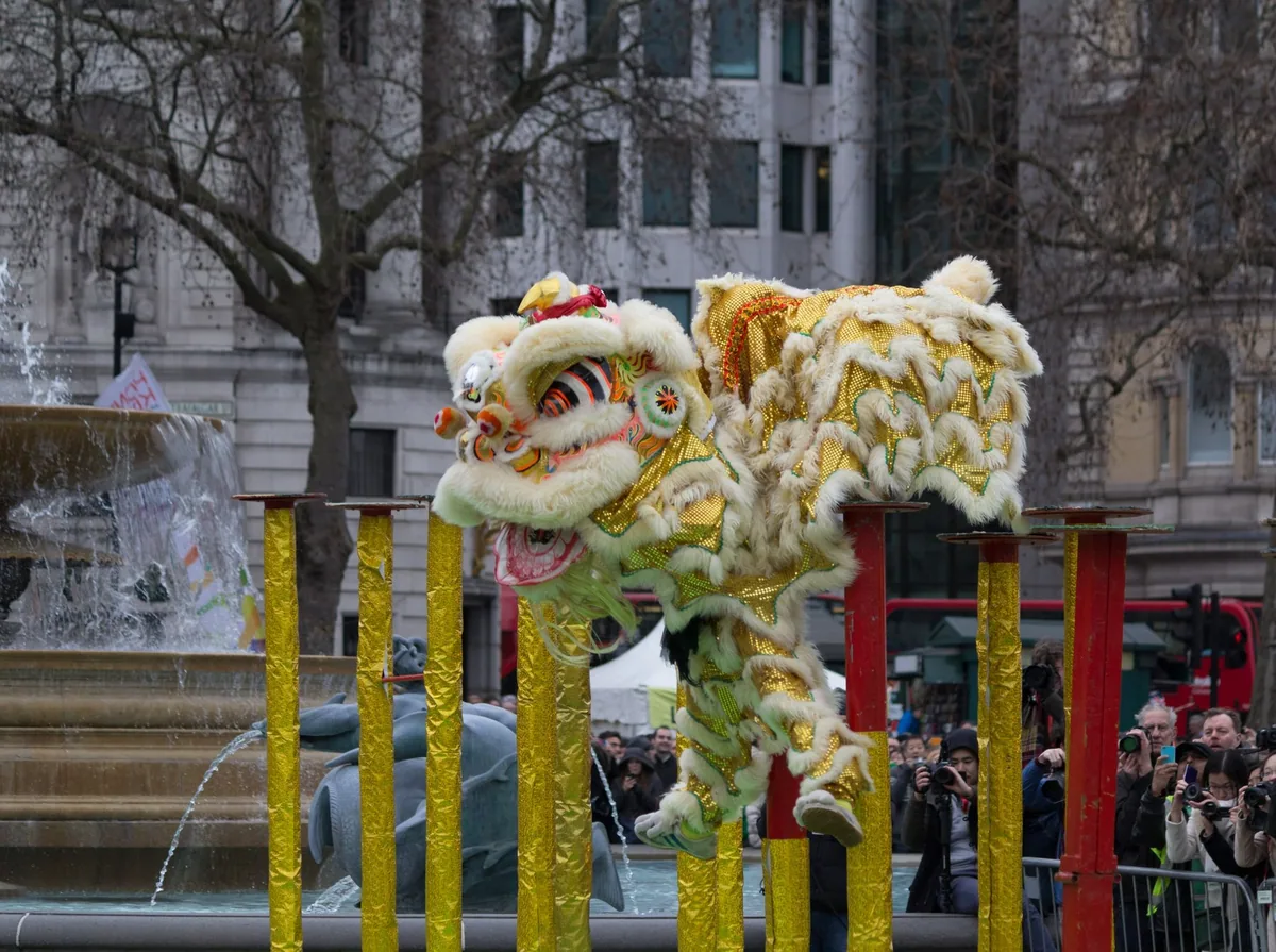 A gold and white Chinese lion dance costume raised on poles in Trafalgar Square, one of the fountains playing behind it