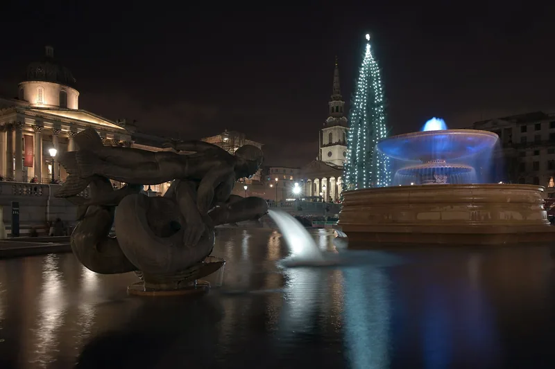 Trafalgar Square at night: the bronze mermaid fountain, a fountain lit blue, and the lit Christmas tree by St Martin-in-the-Fields