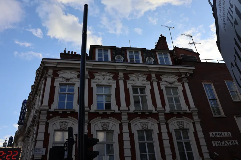 A red-brick and white-stone building on Shaftesbury Avenue with dormer windows in the roof, the Apollo Theatre's blue sign at the left edge