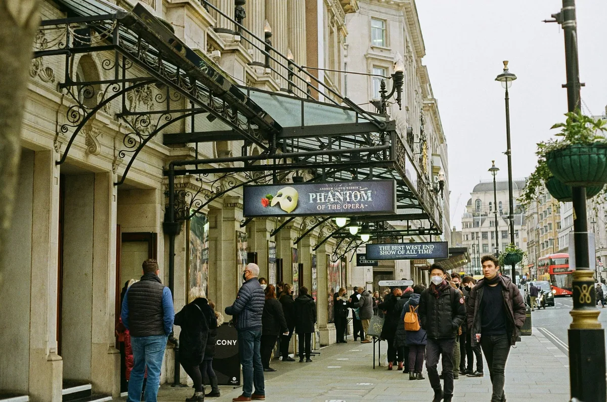 Theatregoers queuing under the glass canopy of Her Majesty's Theatre in London's West End
