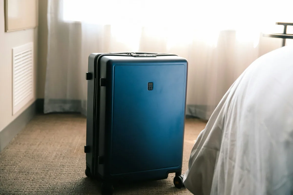 A blue hard-shell suitcase standing on the carpet beside a hotel bed in morning light