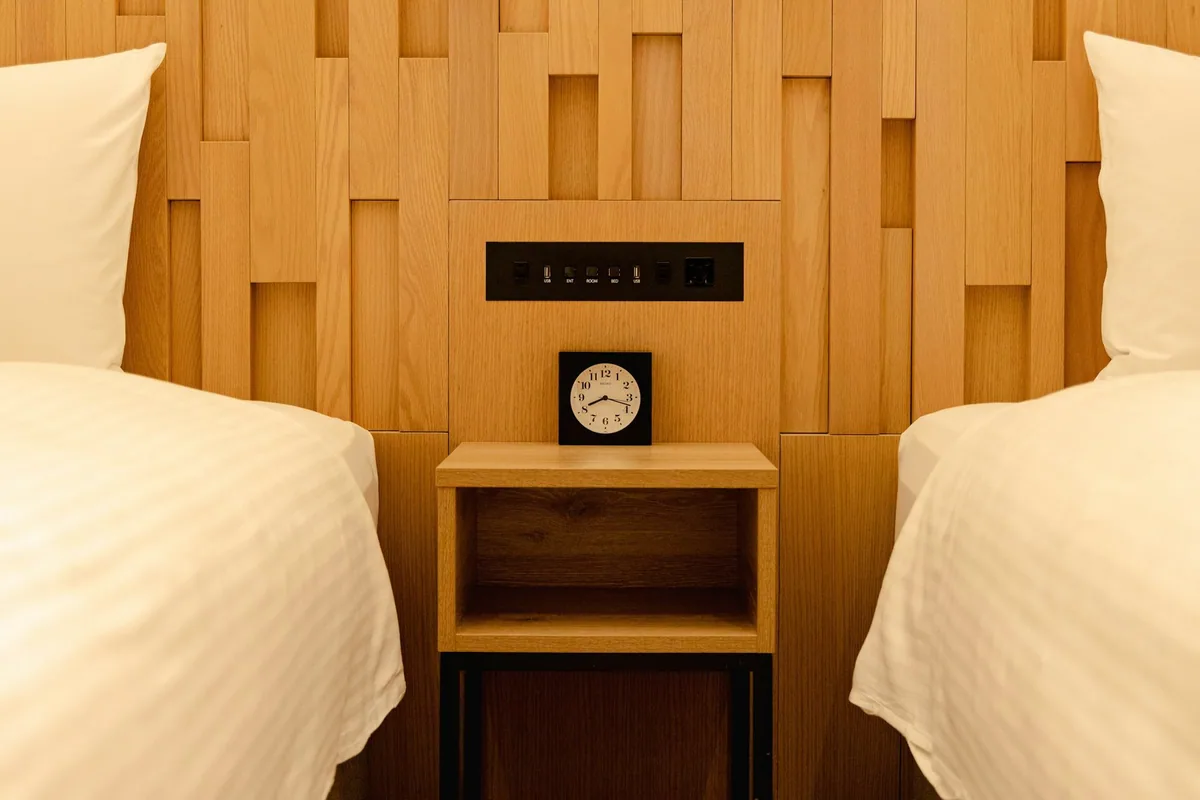 Two single beds either side of a small bedside table with a clock and sockets on a wooden wall
