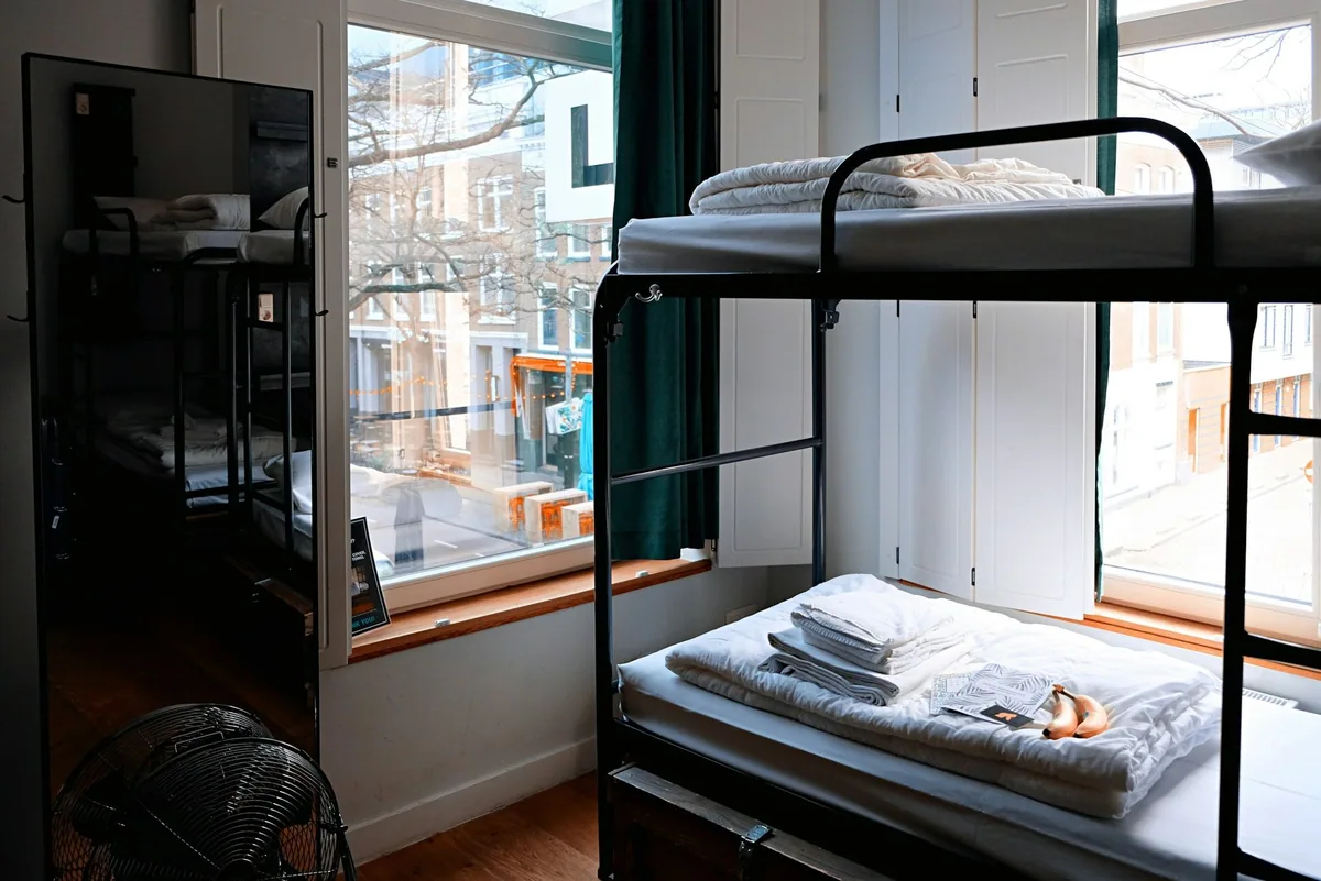 Metal bunk beds with folded towels in a hostel dormitory lit by tall street-facing windows