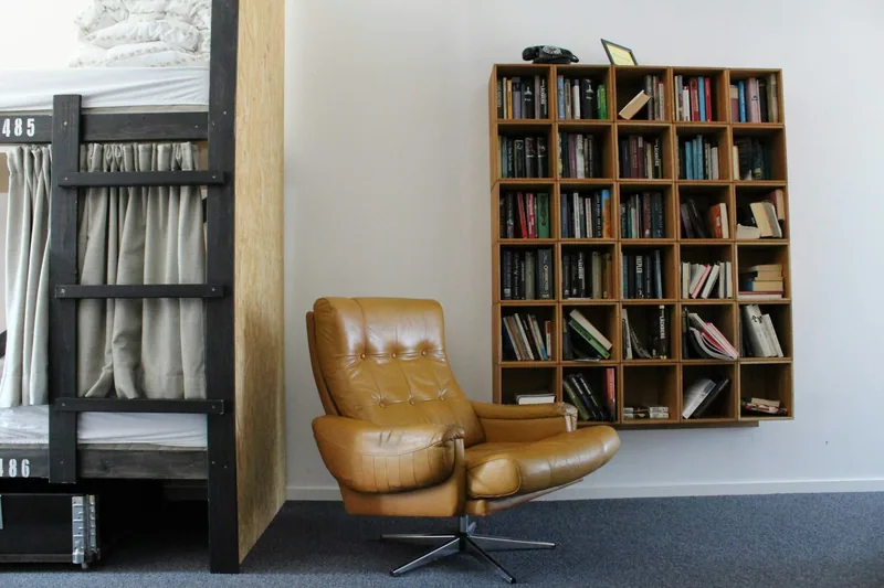 A hostel common area with a book-filled shelf unit and an armchair beside the bunk beds