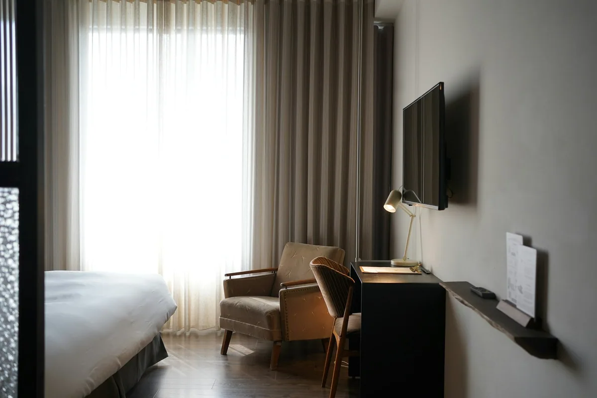 A compact modern hotel room with a desk, armchair and daylight through sheer curtains