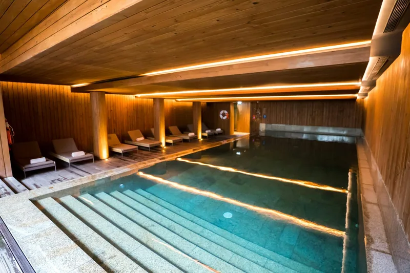 A wood-panelled indoor hotel pool lit by warm strip lighting, with towel-laid loungers on a timber deck beside the water