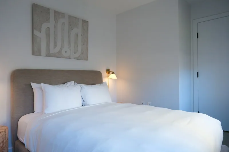 A small white hotel room seen from the door, with a brass wall light over the bed