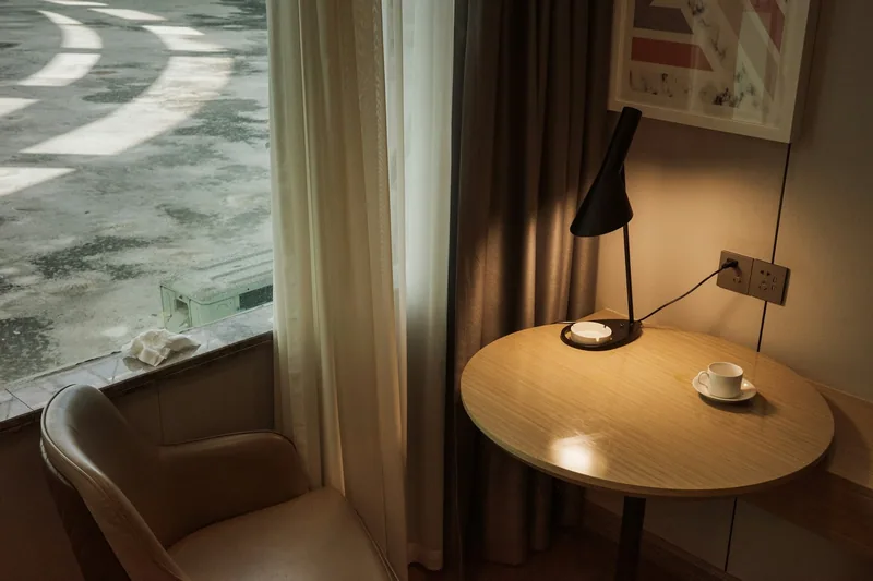 The corner of a hotel room: a round table with a reading lamp and a cup of coffee, an armchair, and a window onto an empty pavement