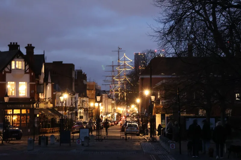A lit street of shops in Greenwich at dusk, with a tall ship rigging strung with lights beyond