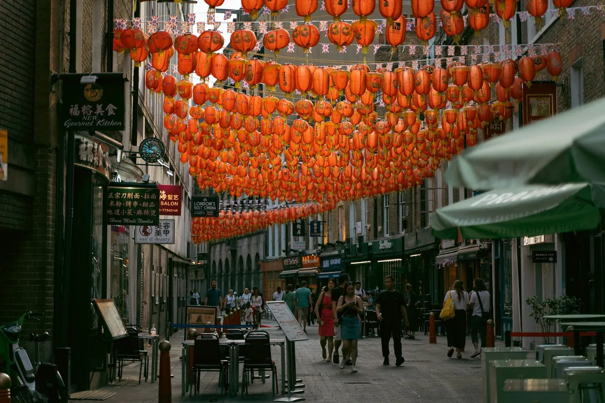 Rows of orange lanterns strung above a Chinatown street in Soho, with cafe tables outside