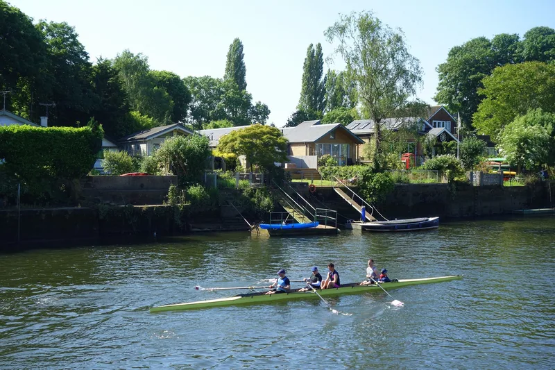A coxed four rowing past the Twickenham bank of the Thames, with low riverside houses and landing stages behind