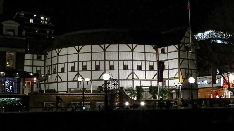 Shakespeare's Globe lit up at night from the riverside walk, its white timber-framed wall and thatched roof above a brick terrace